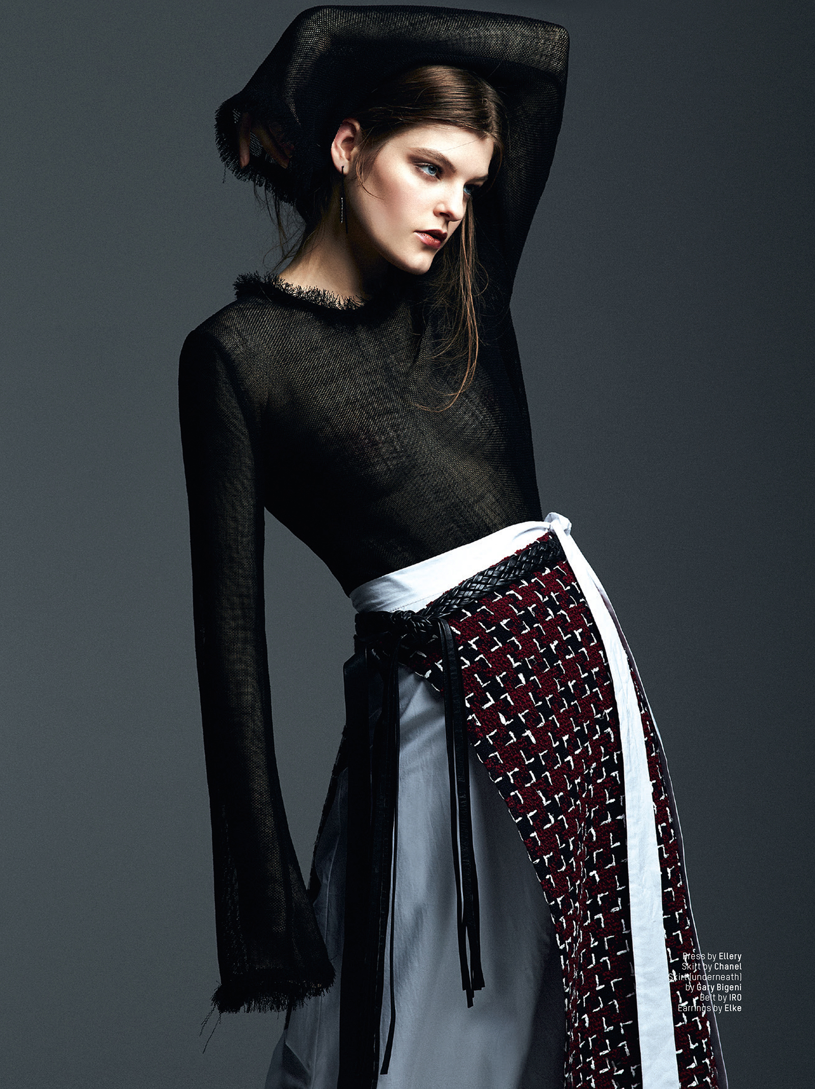 Fashion editorial shot: a brunette adult woman in black top and patterned layered skirt poses against a gray studio backdrop.