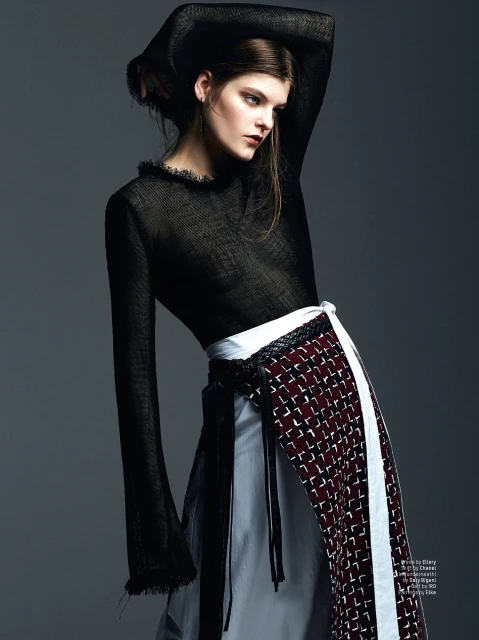 Fashion editorial shot: a brunette adult woman in black top and patterned layered skirt poses against a gray studio backdrop.