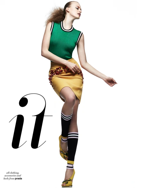 A woman in a green top and yellow skirt poses with an outstretched arm, wearing yellow heels and knee-high socks, set against a white backdrop.