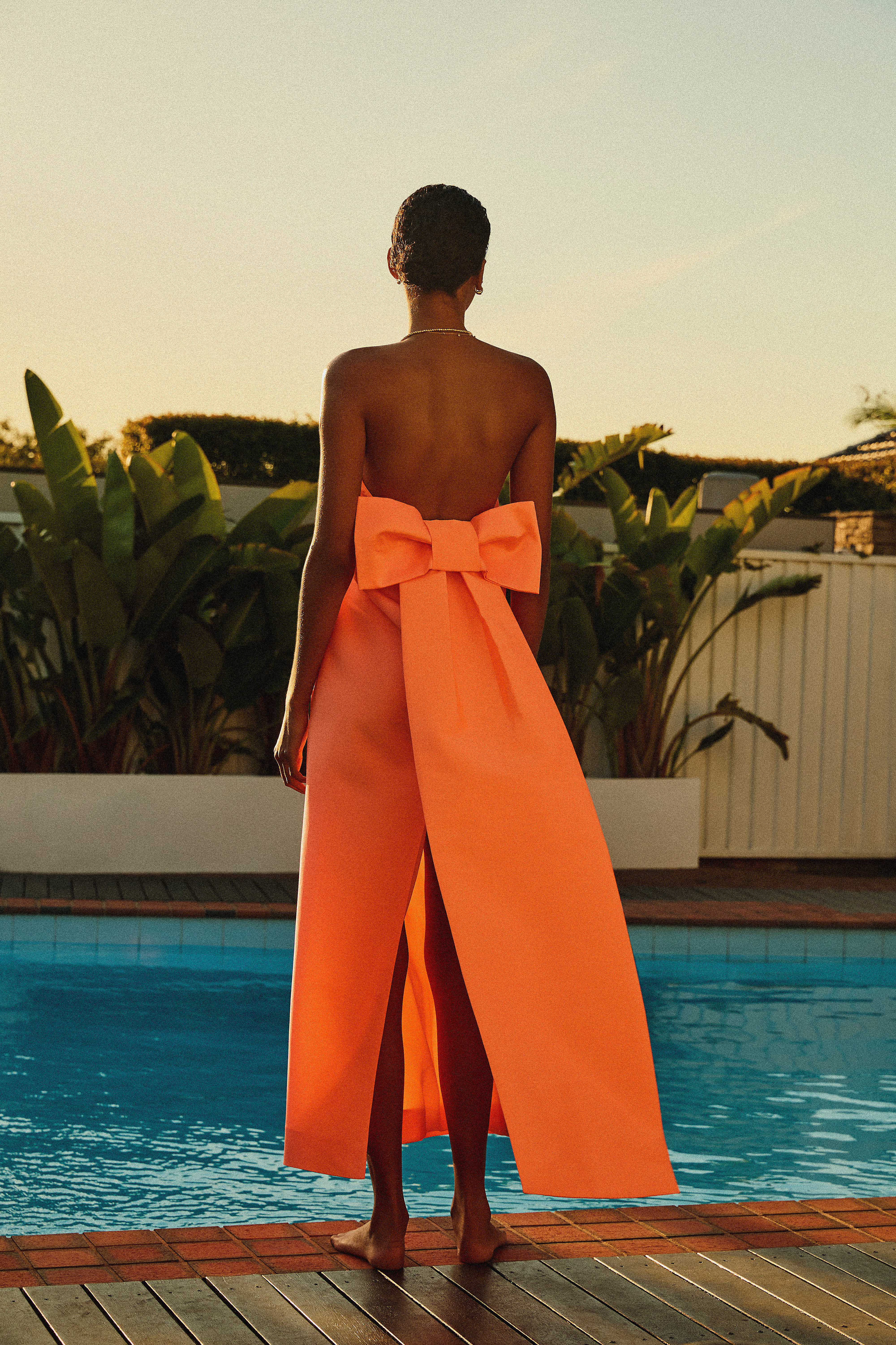 Adult female in coral strapless open-back gown with oversized bow standing near blue swimming pool with lush green plants around perimeter.