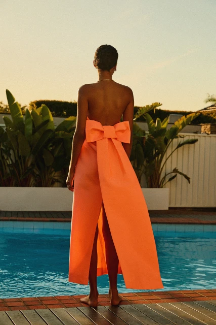 Adult female in coral strapless open-back gown with oversized bow standing near blue swimming pool with lush green plants around perimeter.