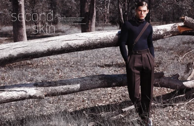 Male model in dark sweater and brown trousers leaning against a large fallen log in a sparse woodland setting by Adam Flipp