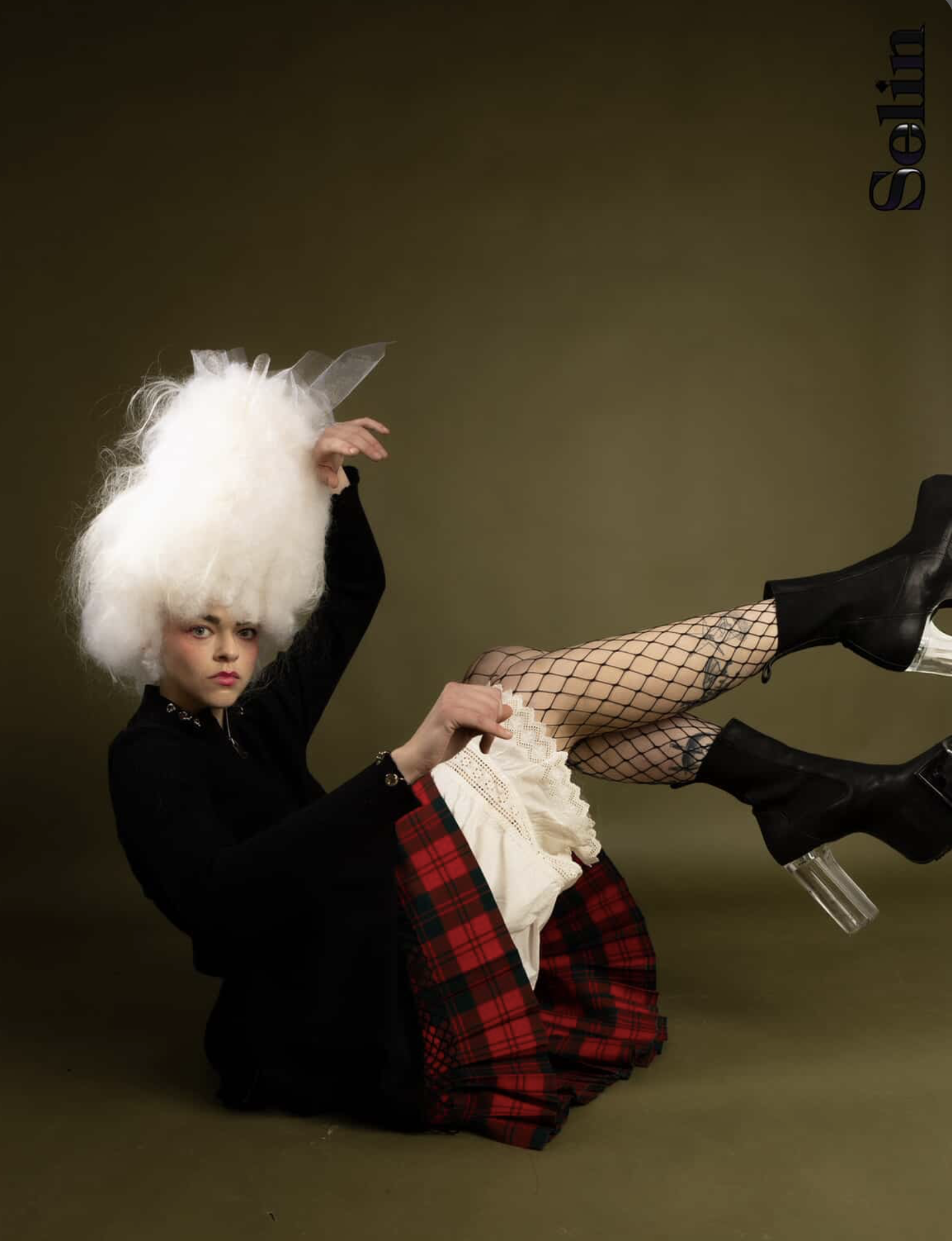 Portrait of a fashion model with unique makeup and hair, wearing black clothing and boots, seated against a neutral olive-green backdrop.