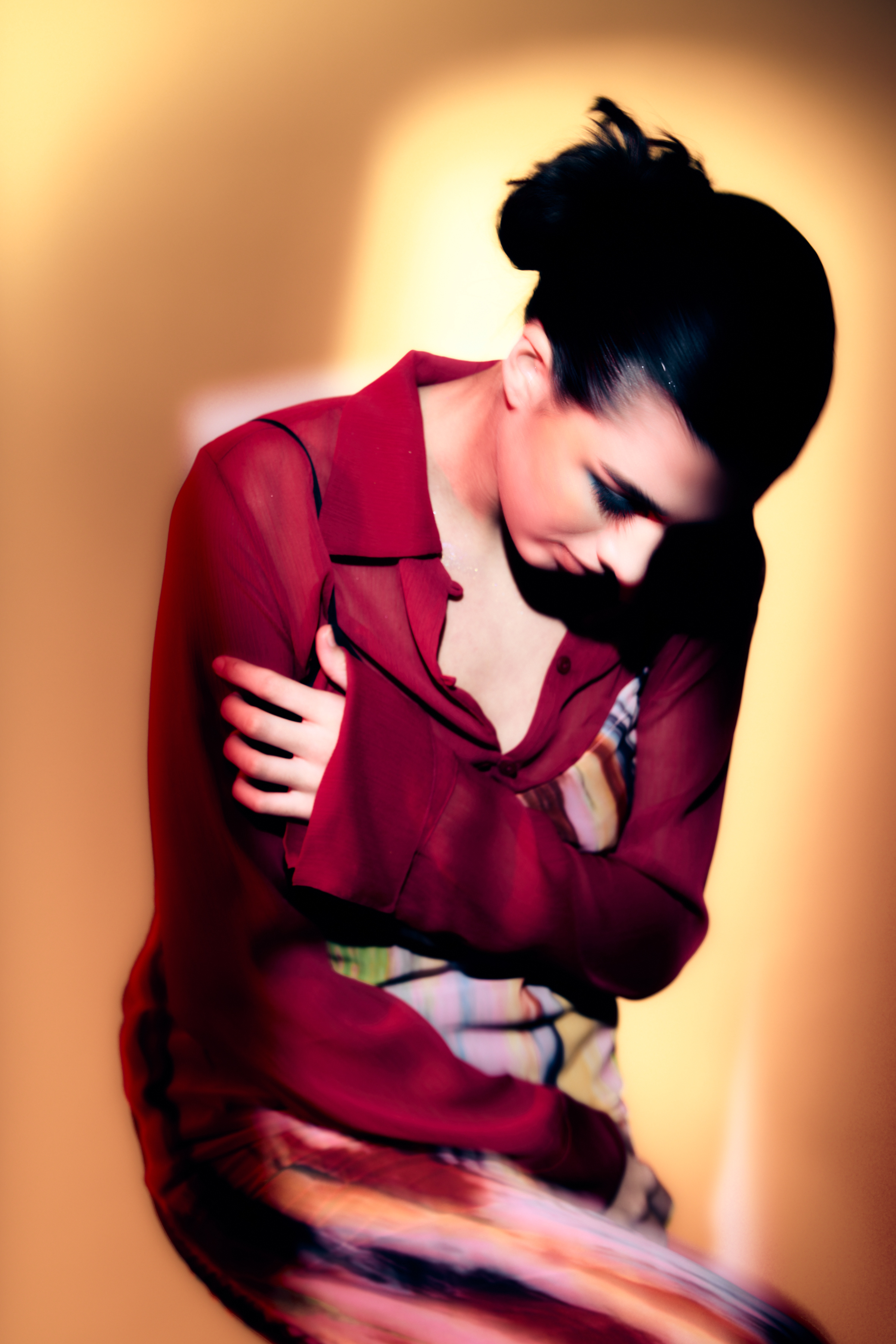 Portrait of a young woman with dark hair wearing a maroon blouse and striped skirt against a peach background in soft studio lighting.