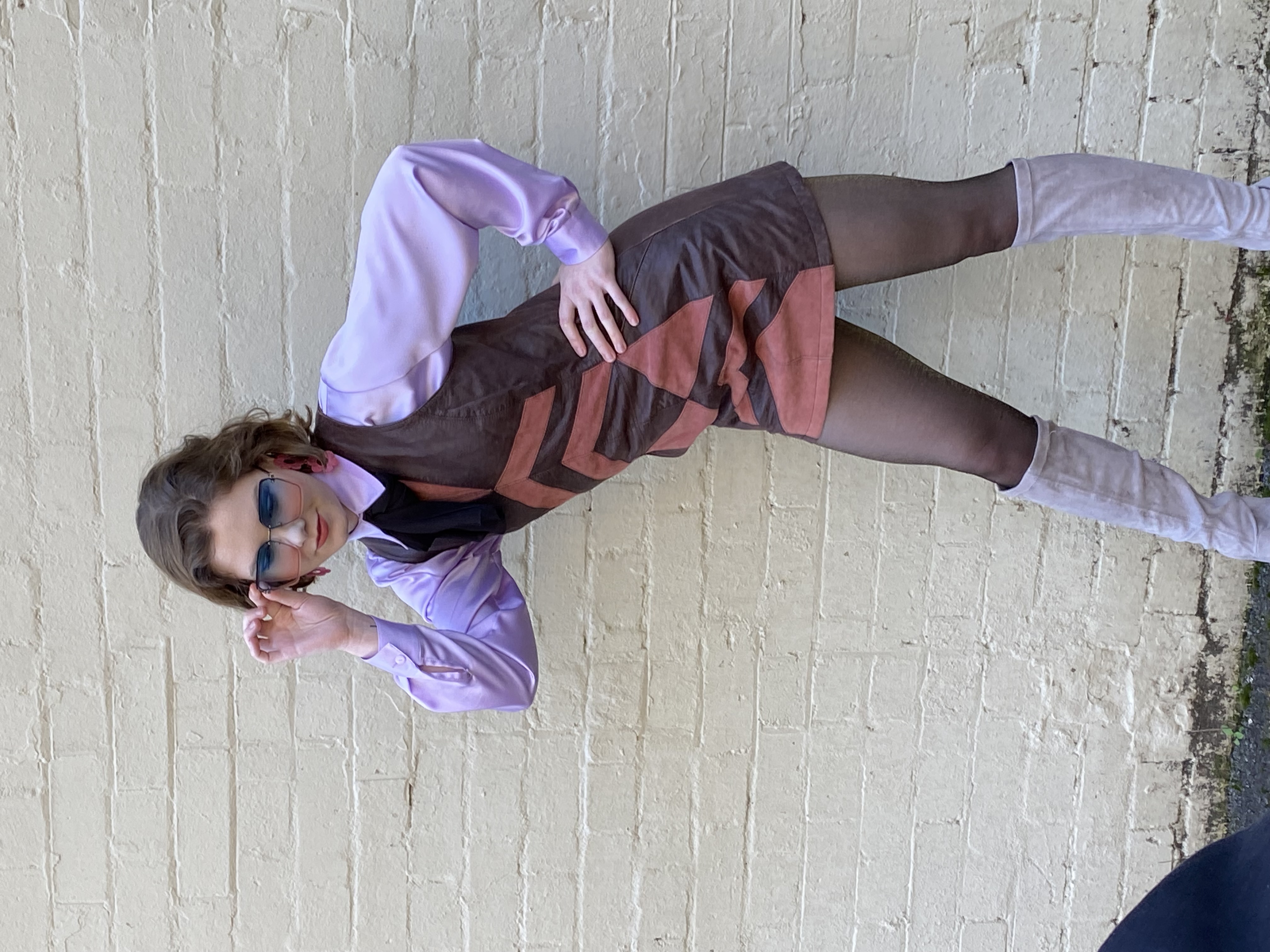 A woman poses in a retro-style brown and orange mini dress with a lilac blouse, tights, and boots against a neutral brick wall.