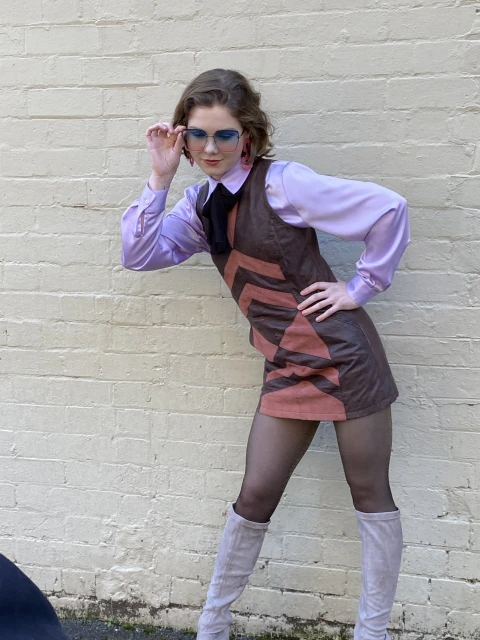A woman poses in a retro-style brown and orange mini dress with a lilac blouse, tights, and boots against a neutral brick wall.