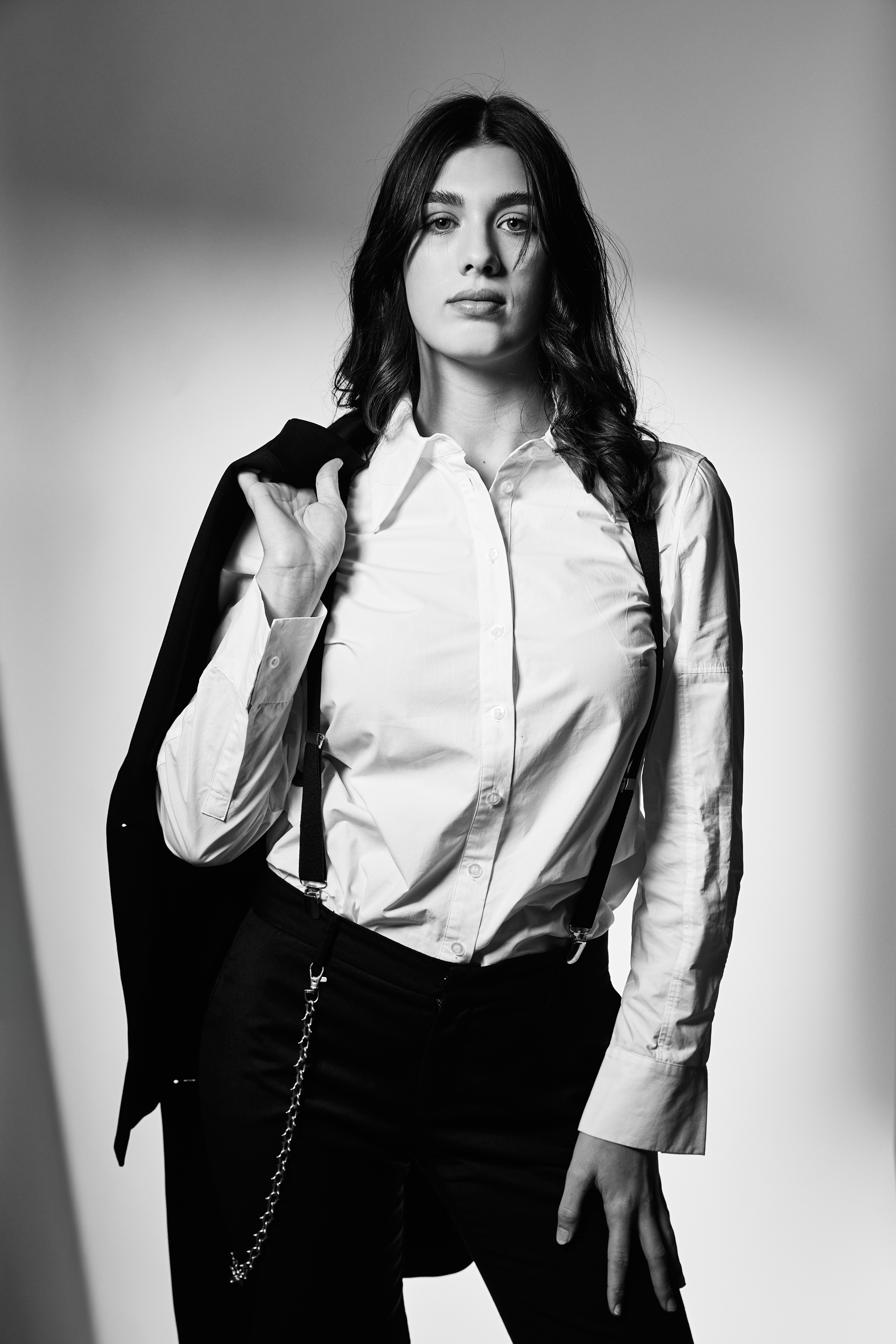 Black and white portrait of a woman in a button-down shirt and suspenders, with a suit jacket draped over her shoulder, indoors.