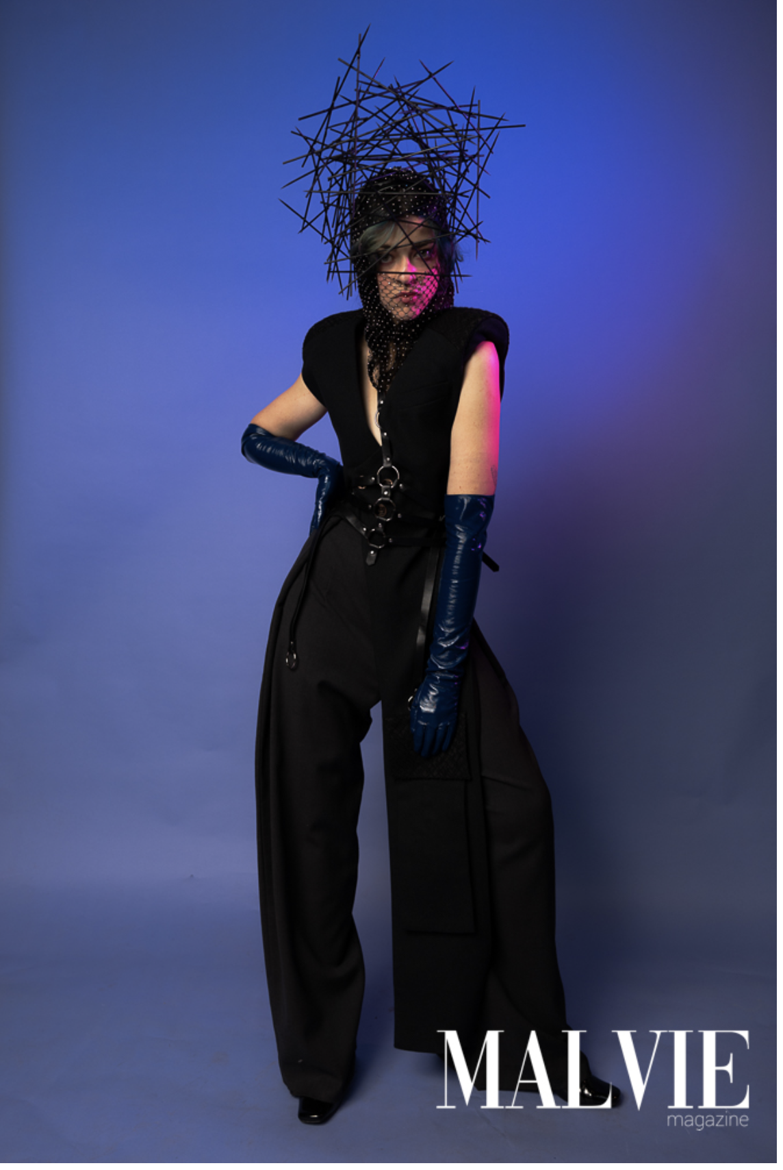 Person in dark clothing and glossy blue gloves poses dramatically against a blue gradient backdrop, wearing artistic headwear for a fashion editorial.
