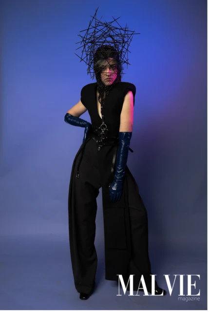 Person in dark clothing and glossy blue gloves poses dramatically against a blue gradient backdrop, wearing artistic headwear for a fashion editorial.