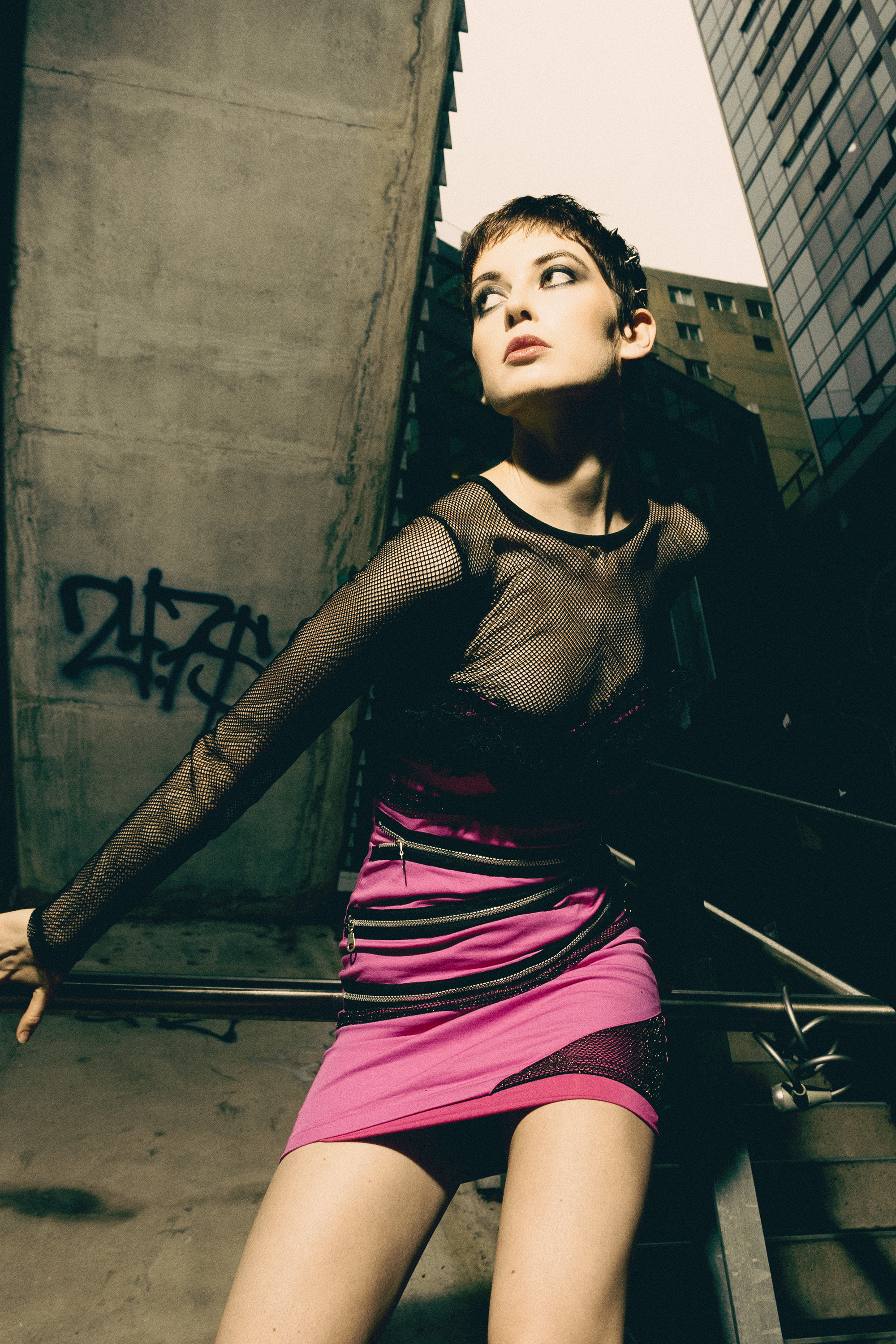 Young woman in pink dress poses in an urban setting with city buildings, wearing a black mesh top, with graffiti on a concrete wall.