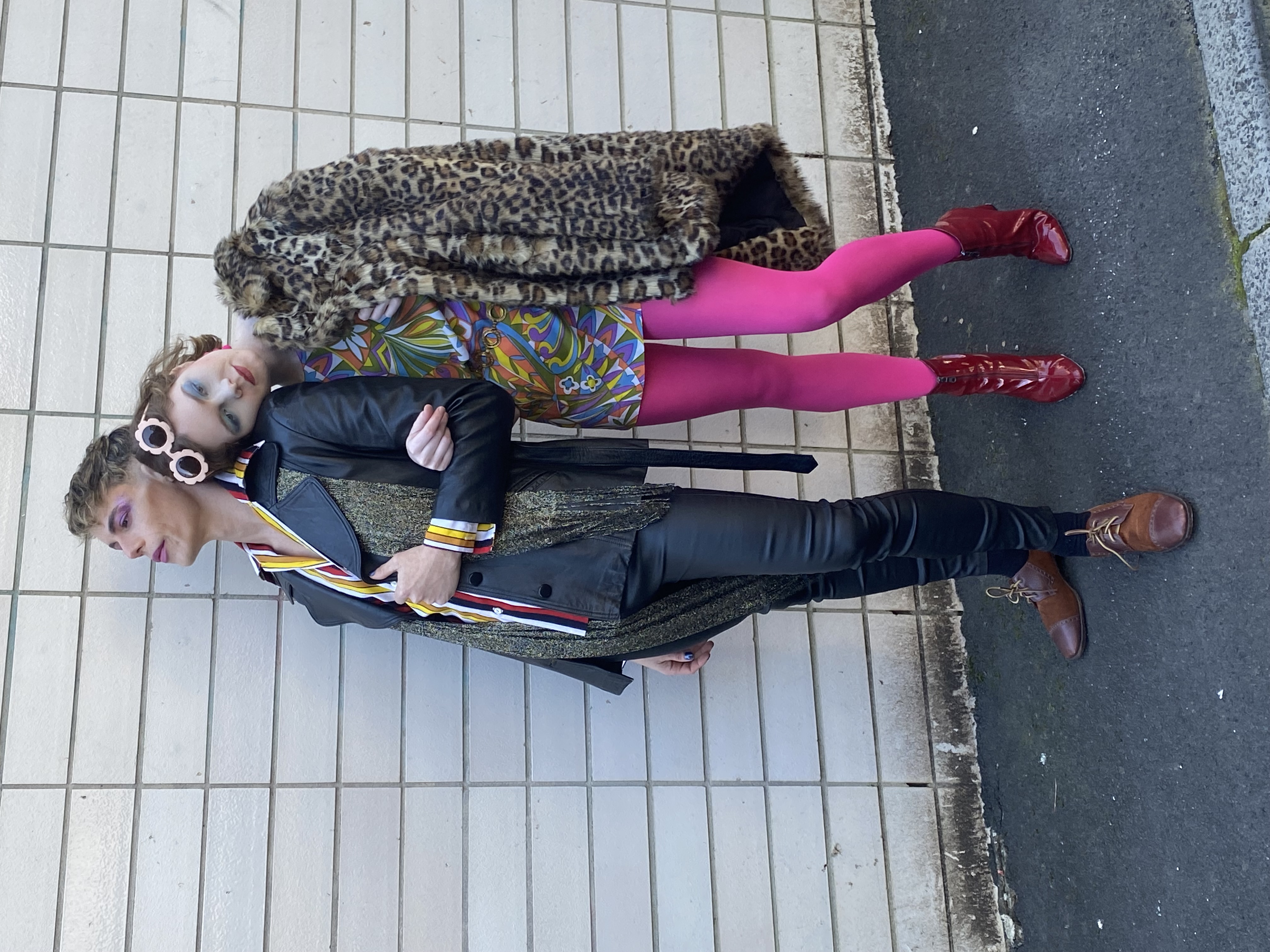 Two people pose outdoors. The person on the left wears a black and gold jacket and pants, while the person on the right wears a patterned dress.