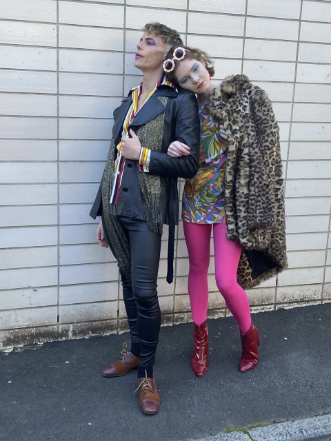 Two people pose outdoors. The person on the left wears a black and gold jacket and pants, while the person on the right wears a patterned dress.