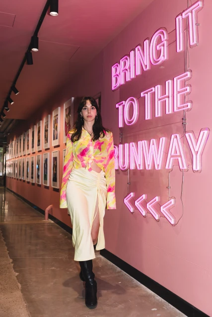 A fashionable young adult woman poses in a stylish yellow and pink outfit in an art gallery with a pink neon sign on the wall.