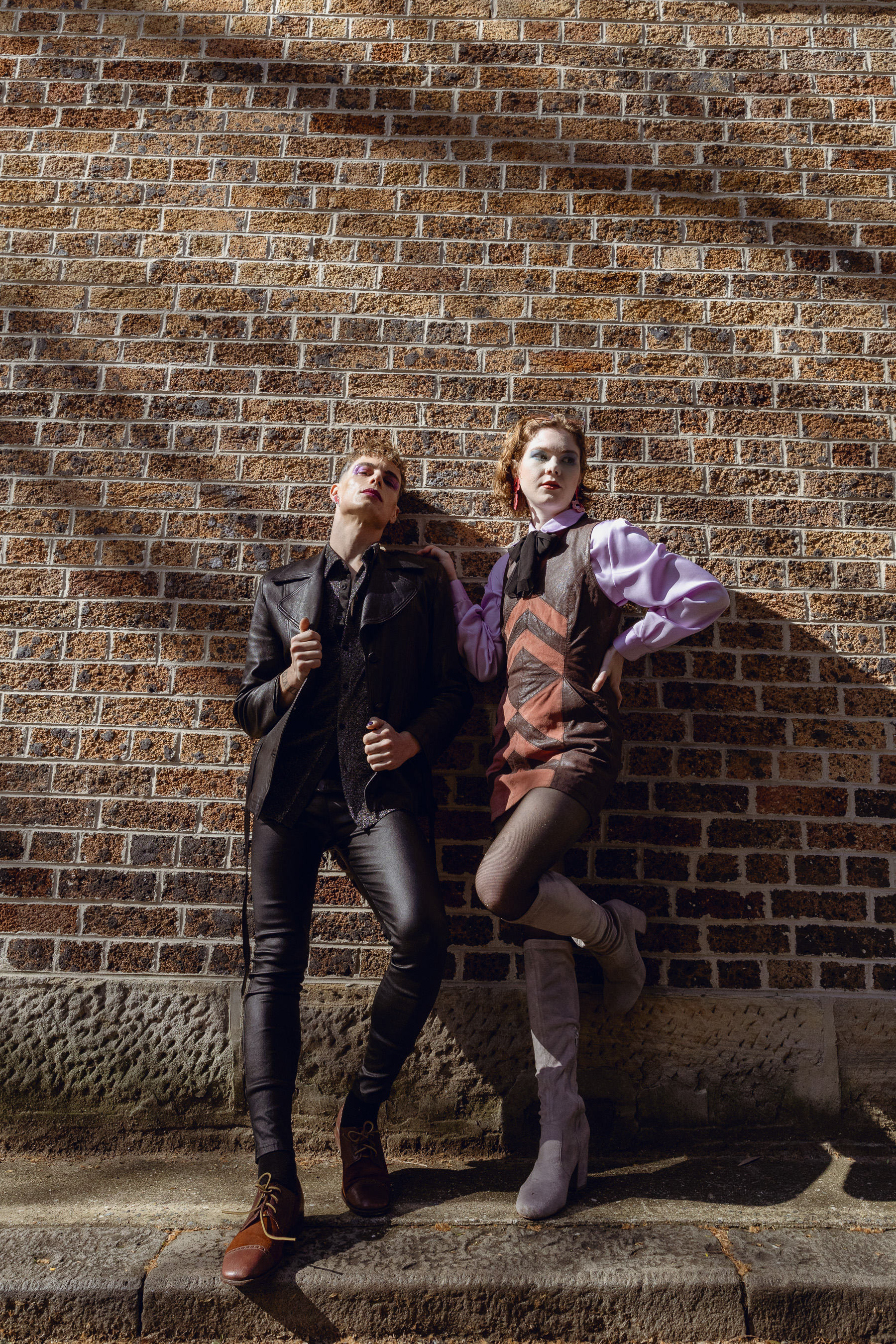 Two fashion models pose against a brick wall in sunlight. The model on the left wears a black leather jacket and pants, while the other wears a dress and boots.