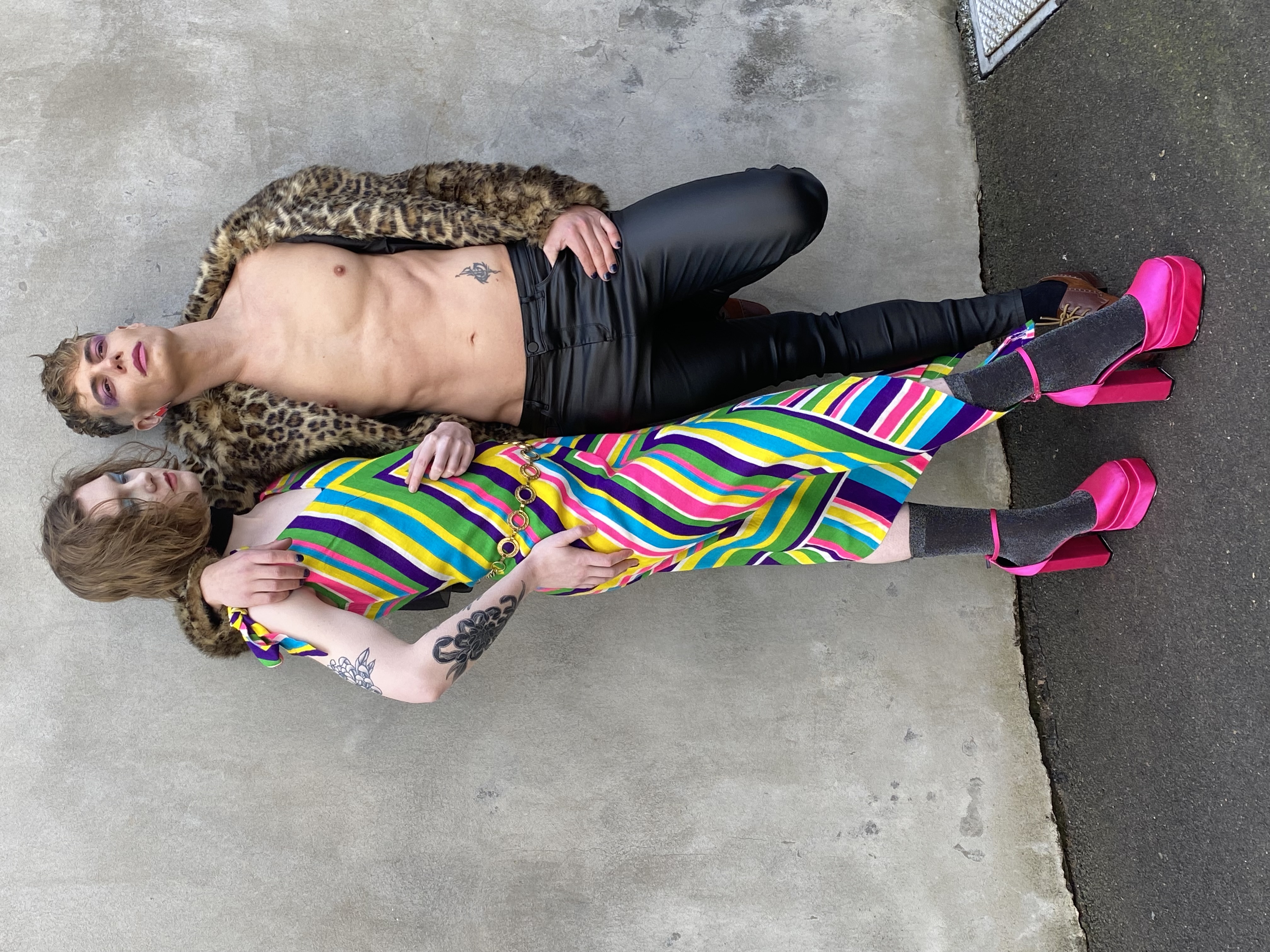 Two people pose in colorful clothing. One wears a striped dress and pink platforms, the other a leopard jacket and leather pants.