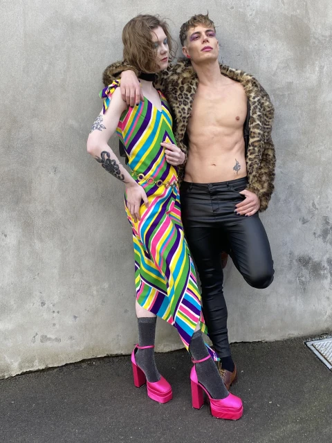 Two people pose in colorful clothing. One wears a striped dress and pink platforms, the other a leopard jacket and leather pants.