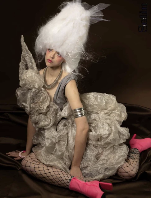 A woman poses wearing a large white wig, sculptural gray dress, pink ankle boots, and fishnet stockings against a brown backdrop.