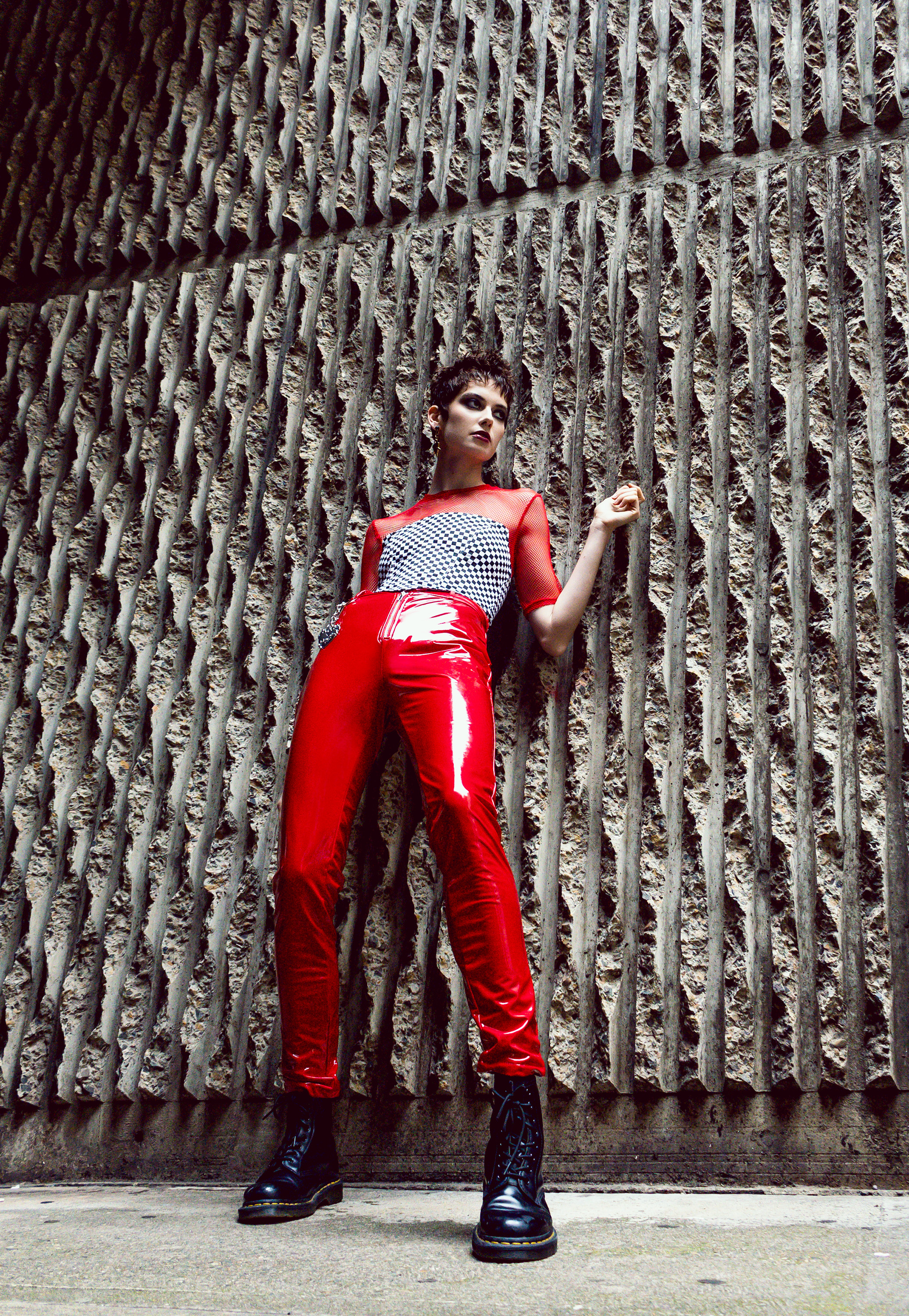 A person with short dark hair poses in red pants and black boots against a patterned concrete wall outdoors in a full shot.
