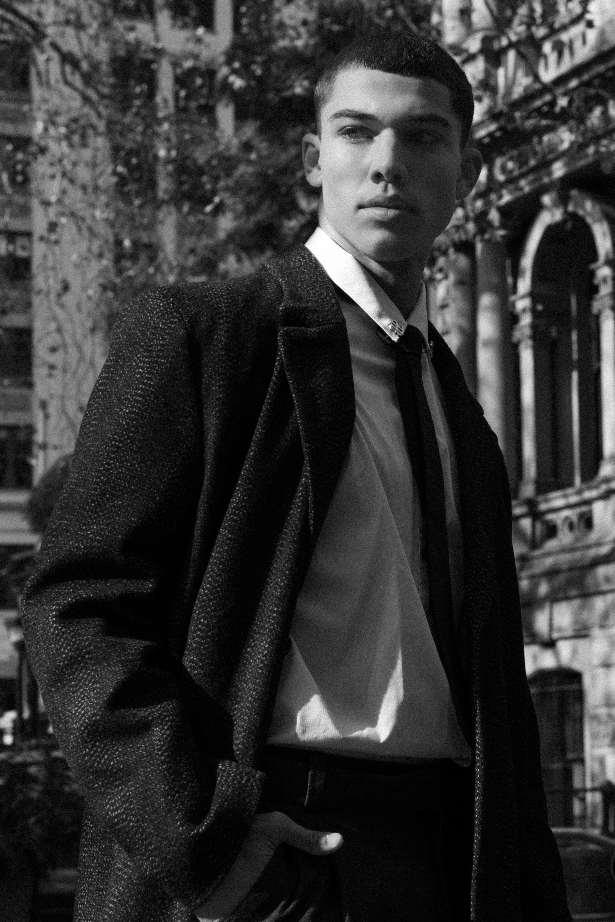 Monochrome portrait of a stylish young man in a suit and tie, standing outdoors with classic architecture and trees visible in the background.
