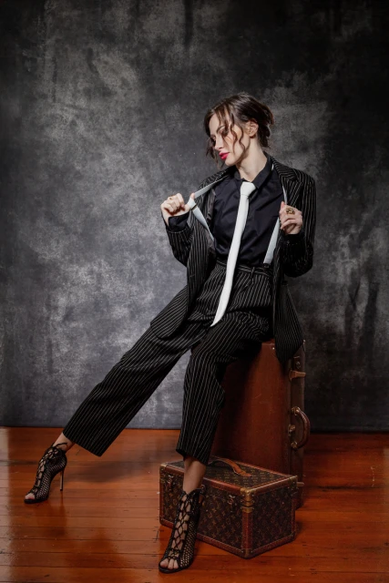 A woman in a pinstripe suit, black shirt, and tie sits on a vintage suitcase in a studio setting, showcasing a blend of fashion and retro style.