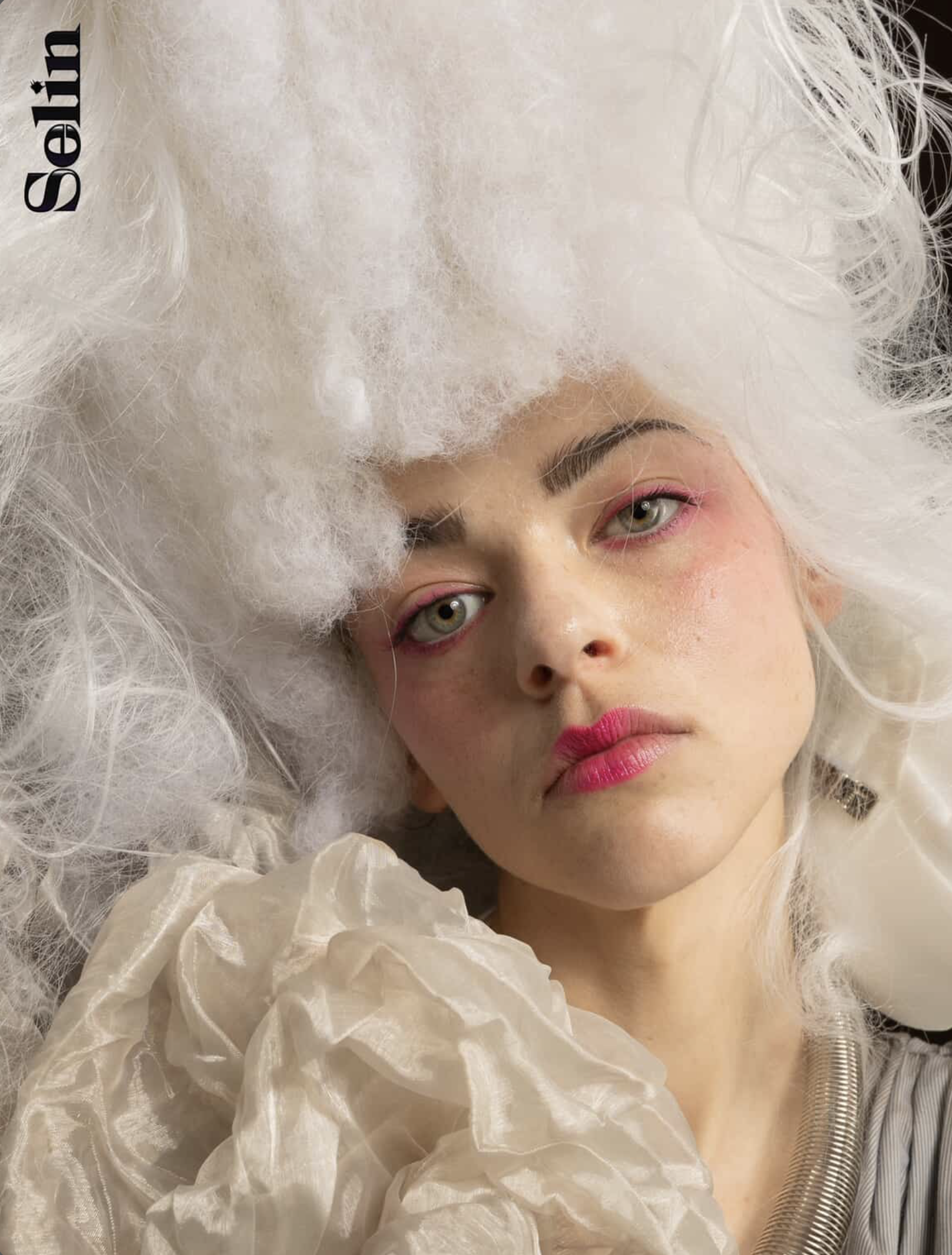 Portrait of a young woman with pink makeup and a large white wig-like structure, gazing directly at the viewer in a studio setting.