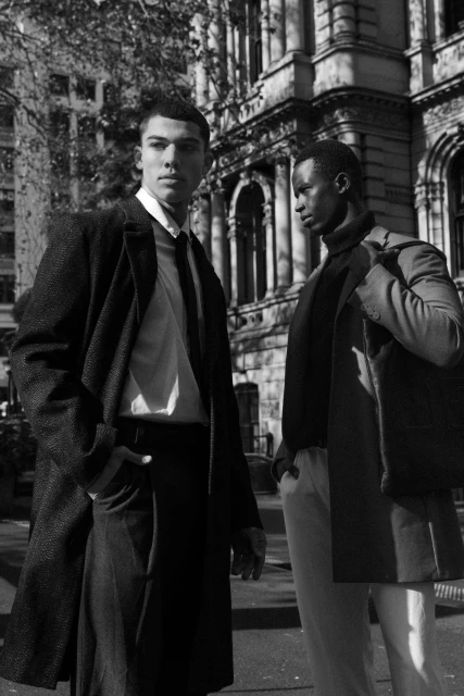 Two male fashion models in coats and suits stand outside an urban building in black and white, exuding a sophisticated, stylish vibe.