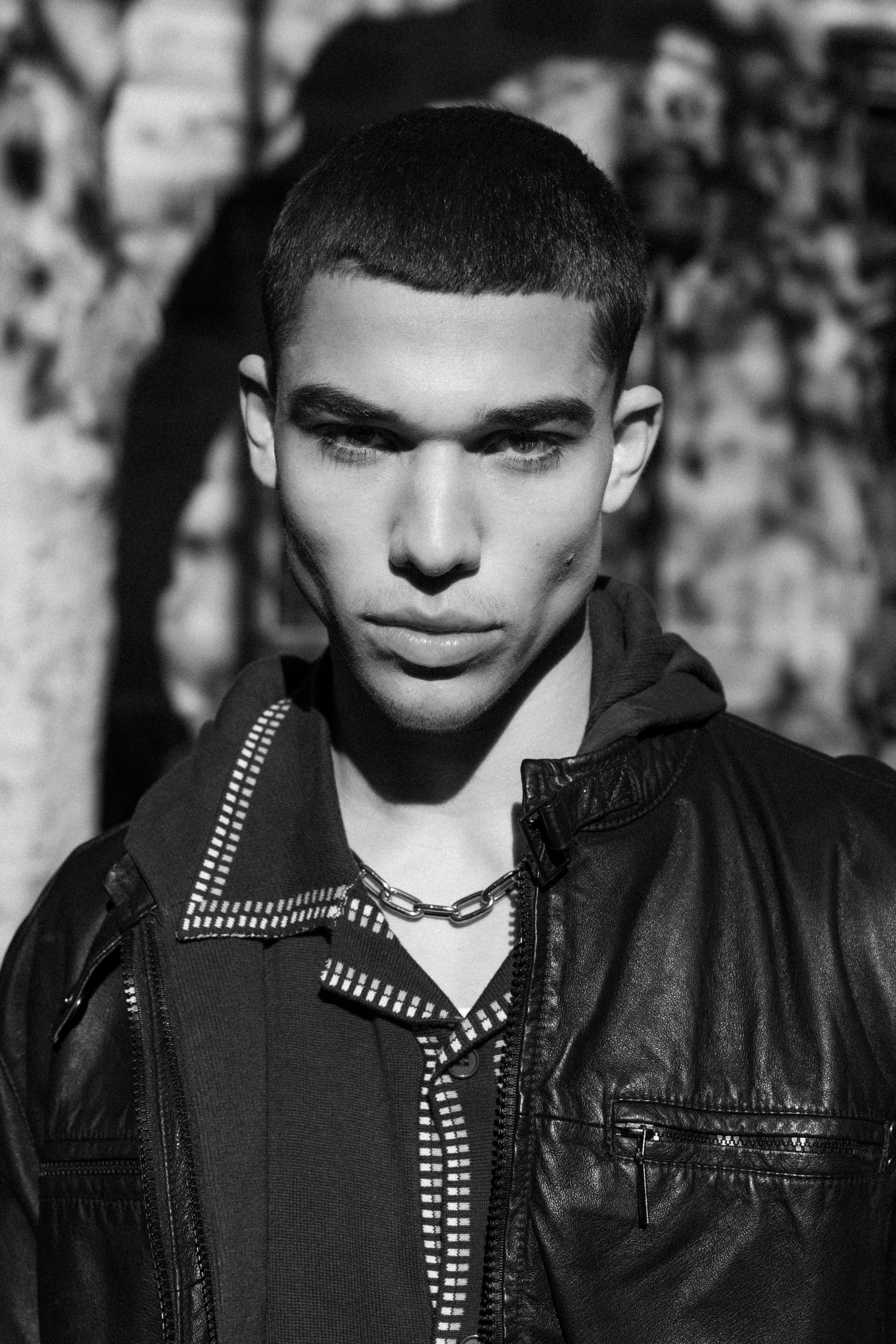 Monochrome portrait of a young adult male with a serious expression, wearing a leather jacket and silver chain necklace, against an outdoor backdrop.