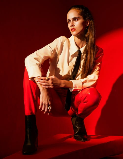 Fashionable woman in cream blouse, black tie, and red pants poses in a photo studio with red walls, wearing black boots and bold red lipstick.