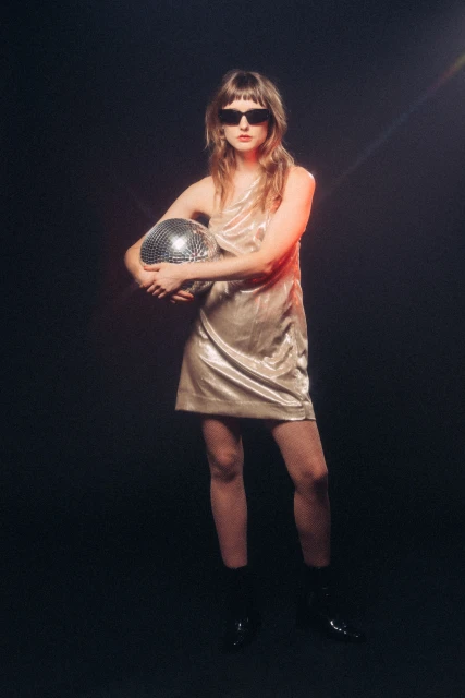A young woman poses in a shimmering champagne dress and black sunglasses, holding a disco ball in front of a dark background.