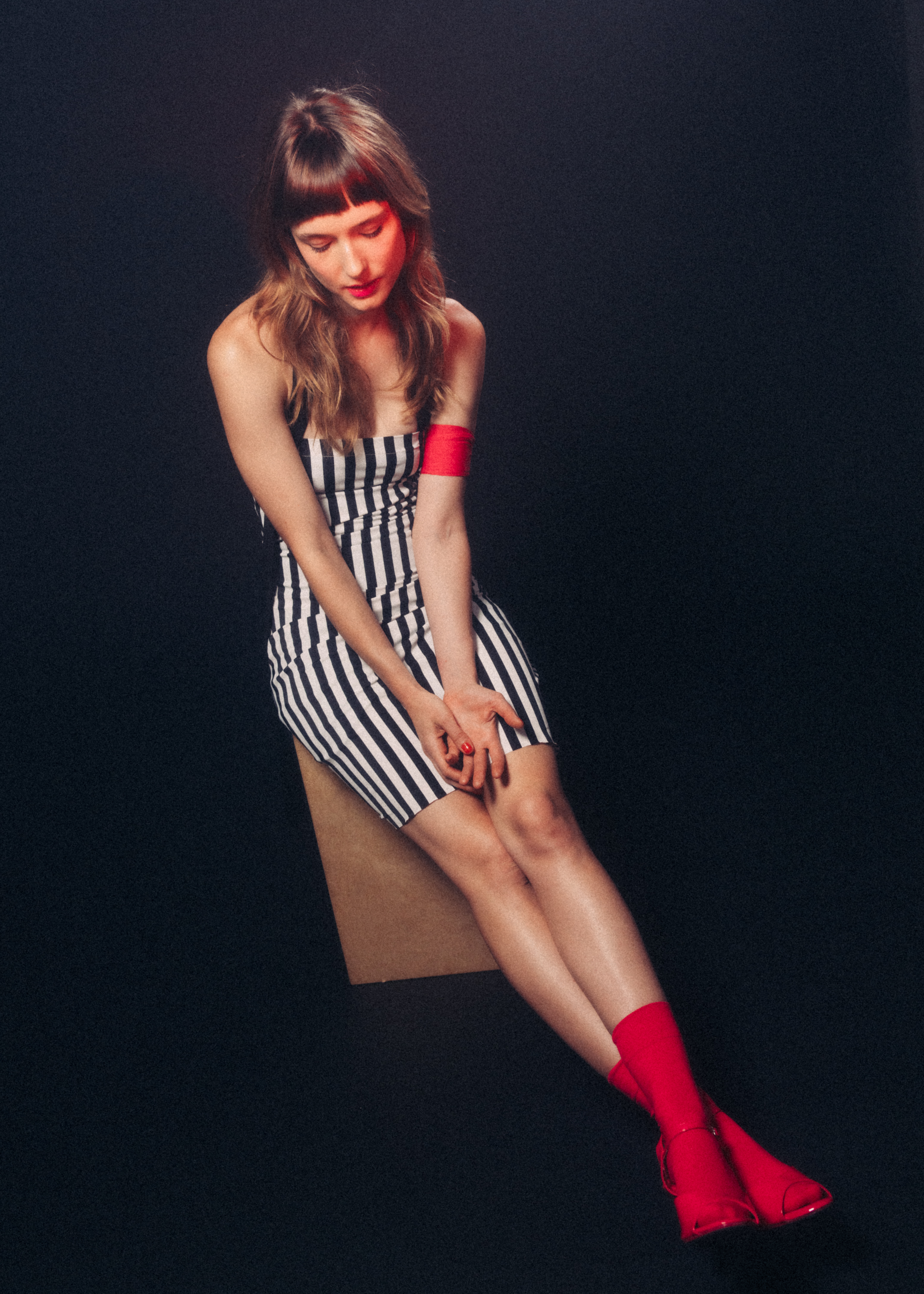 Striking studio portrait of a woman in a striped dress, seated and gazing down, enhanced by red lighting and bold red boots against a black background.