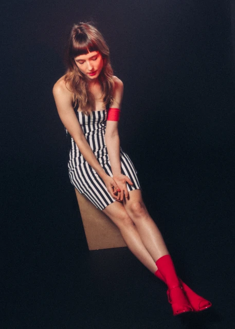 Striking studio portrait of a woman in a striped dress, seated and gazing down, enhanced by red lighting and bold red boots against a black background.