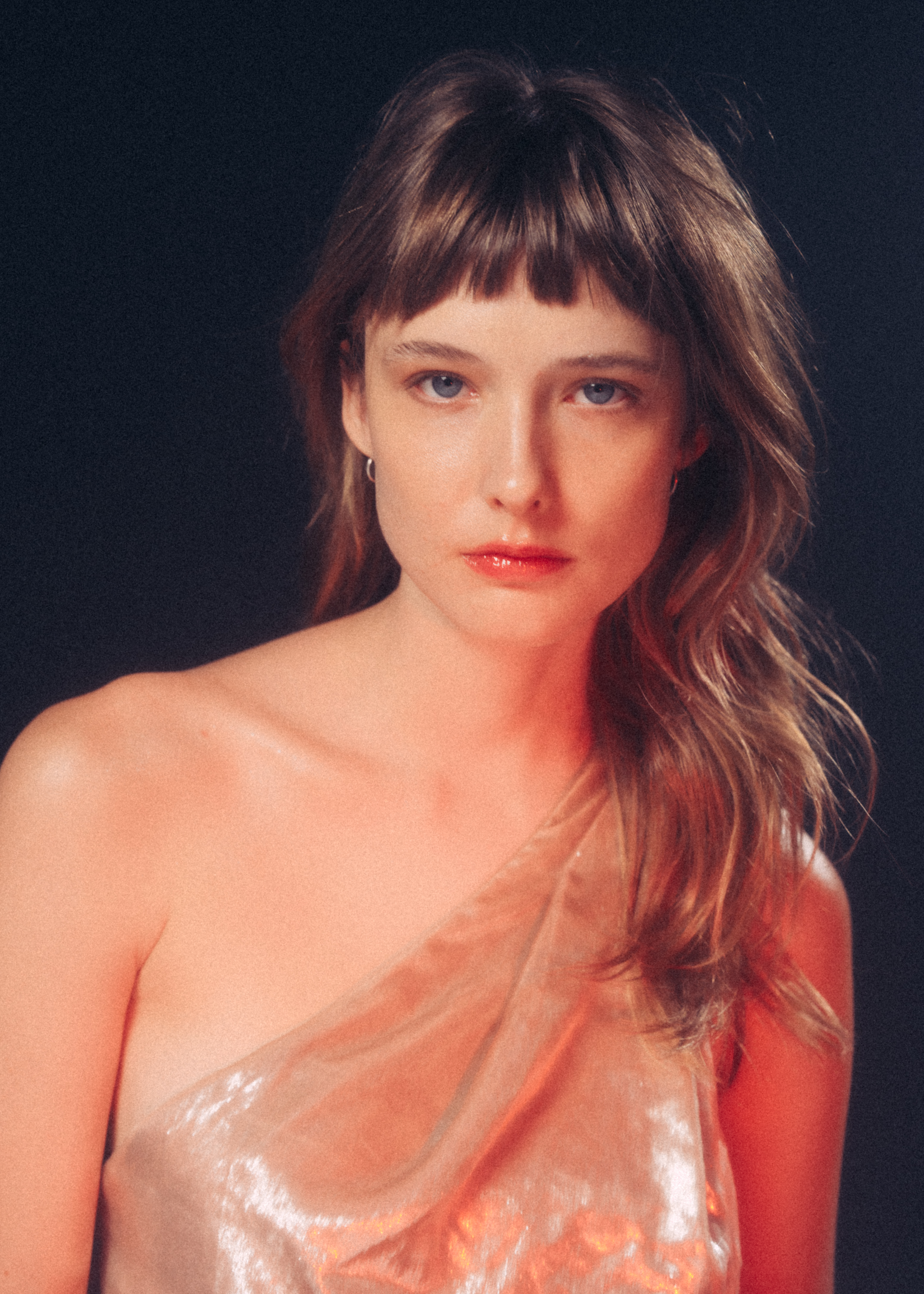 Portrait of a young woman with bangs wearing a shimmering, one-shoulder dress against a dark background in a beauty-style photograph.