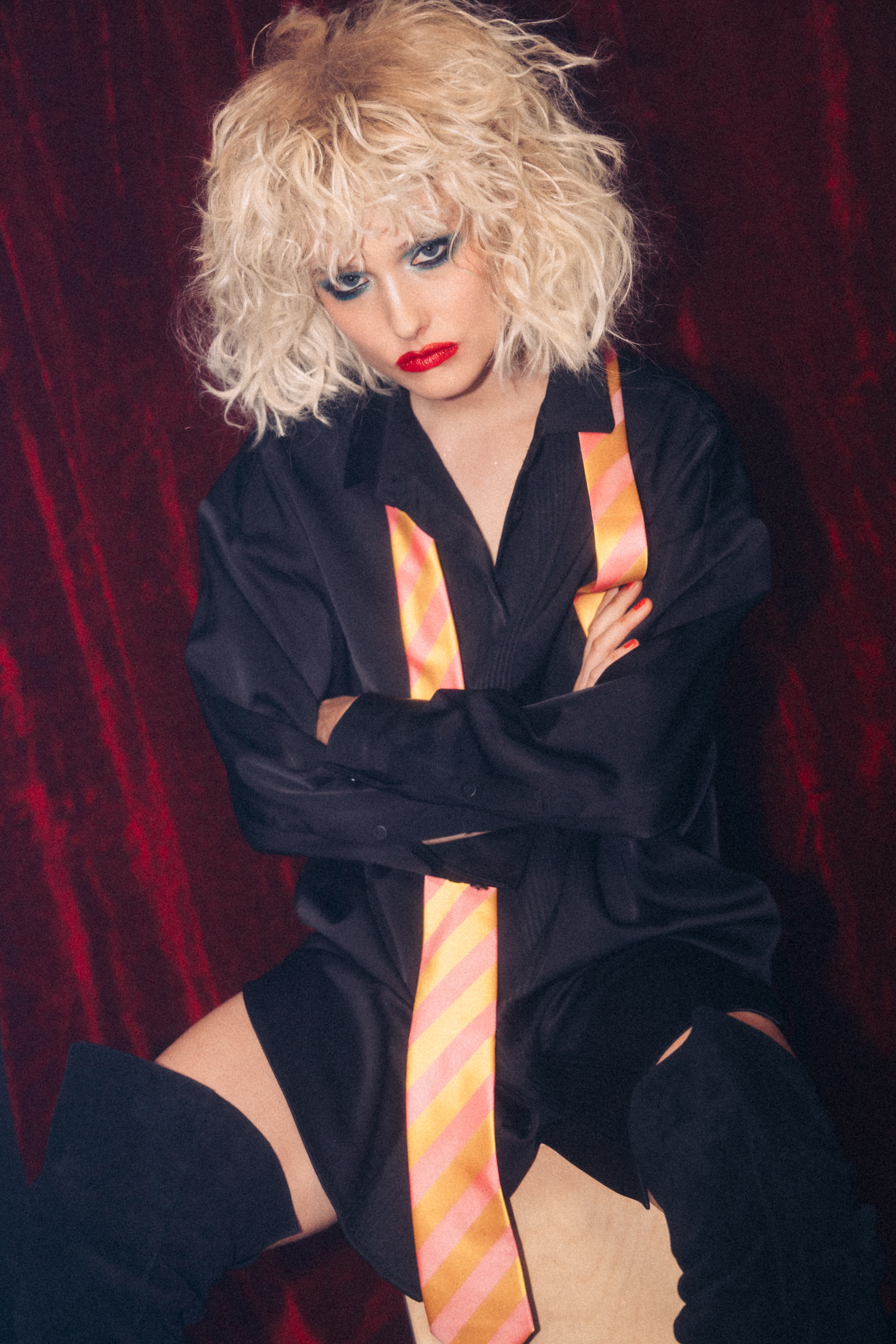 Model with blonde curly hair wearing a black shirt, red lipstick, and striped tie against a dark red velvet background.