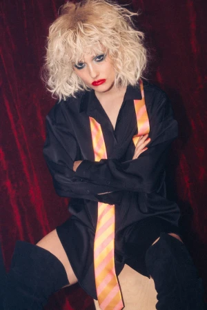 Model with blonde curly hair wearing a black shirt, red lipstick, and striped tie against a dark red velvet background.