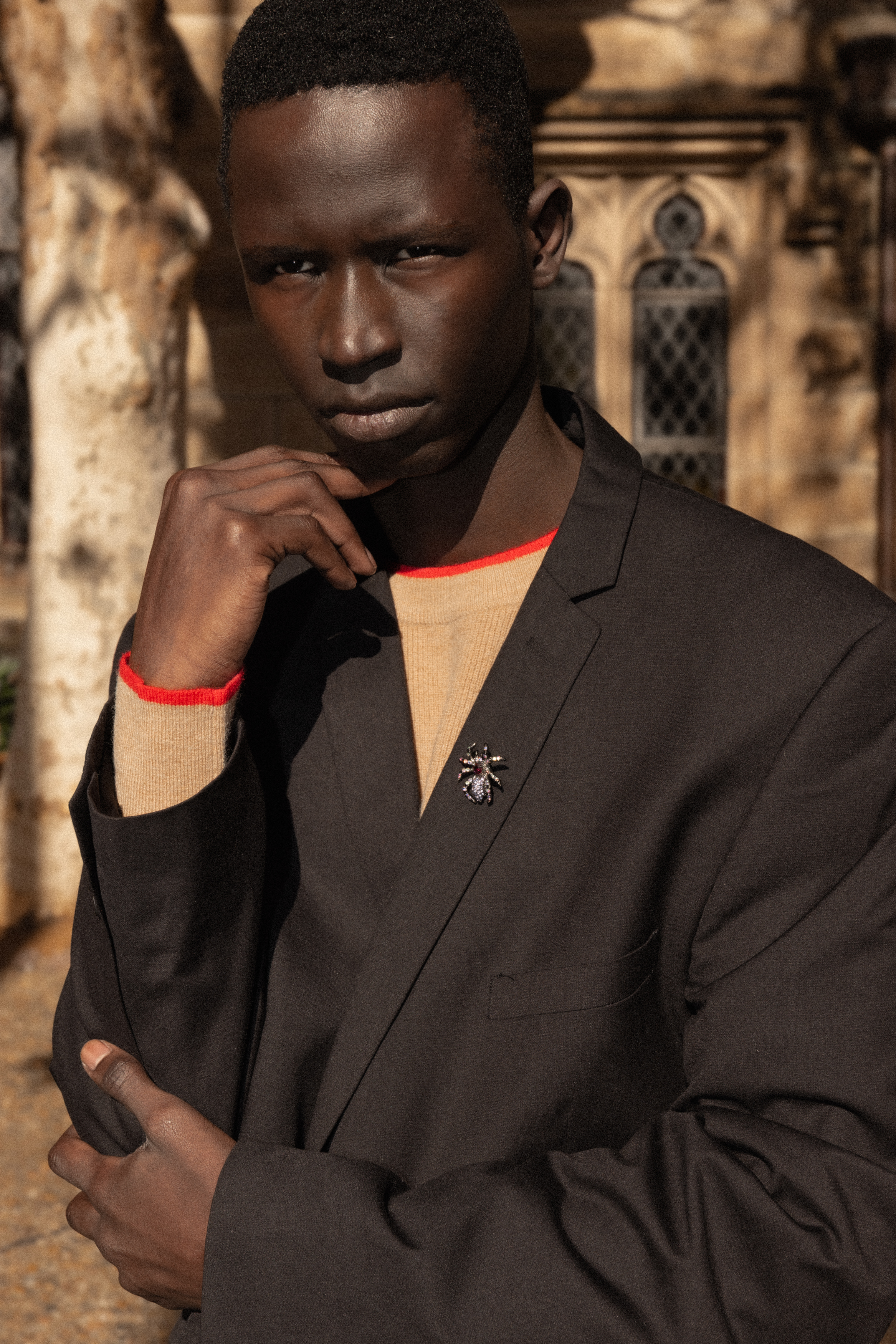 Portrait of an adult male of African descent in a black suit jacket and tan shirt, posed outdoors with natural light and a stone building background.