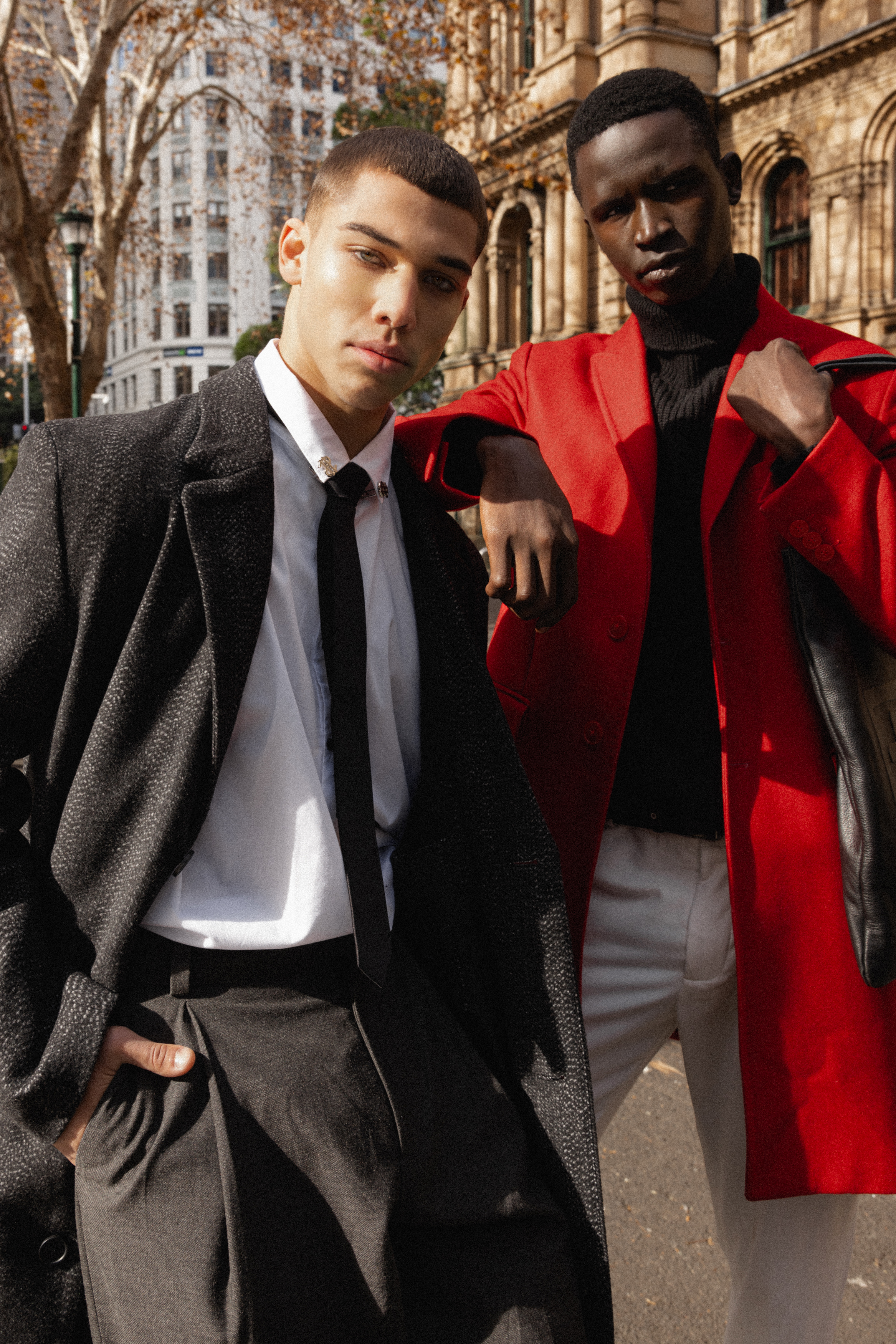 Two stylish men in coats pose in an urban setting. One man wears a red coat, the other a black coat and tie, creating a striking fashion statement.