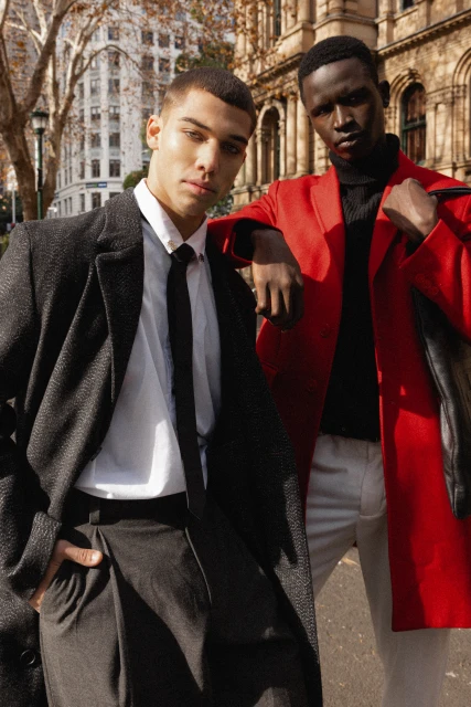 Two stylish men in coats pose in an urban setting. One man wears a red coat, the other a black coat and tie, creating a striking fashion statement.