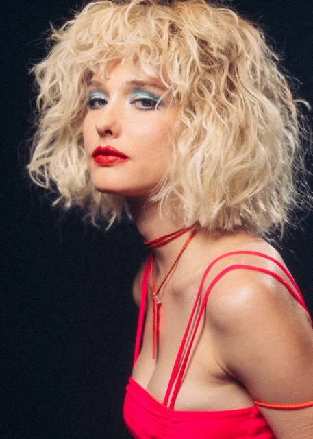 Portrait of a woman with curly blonde hair, bright blue eyeshadow, and red lipstick in front of a dark black background. 