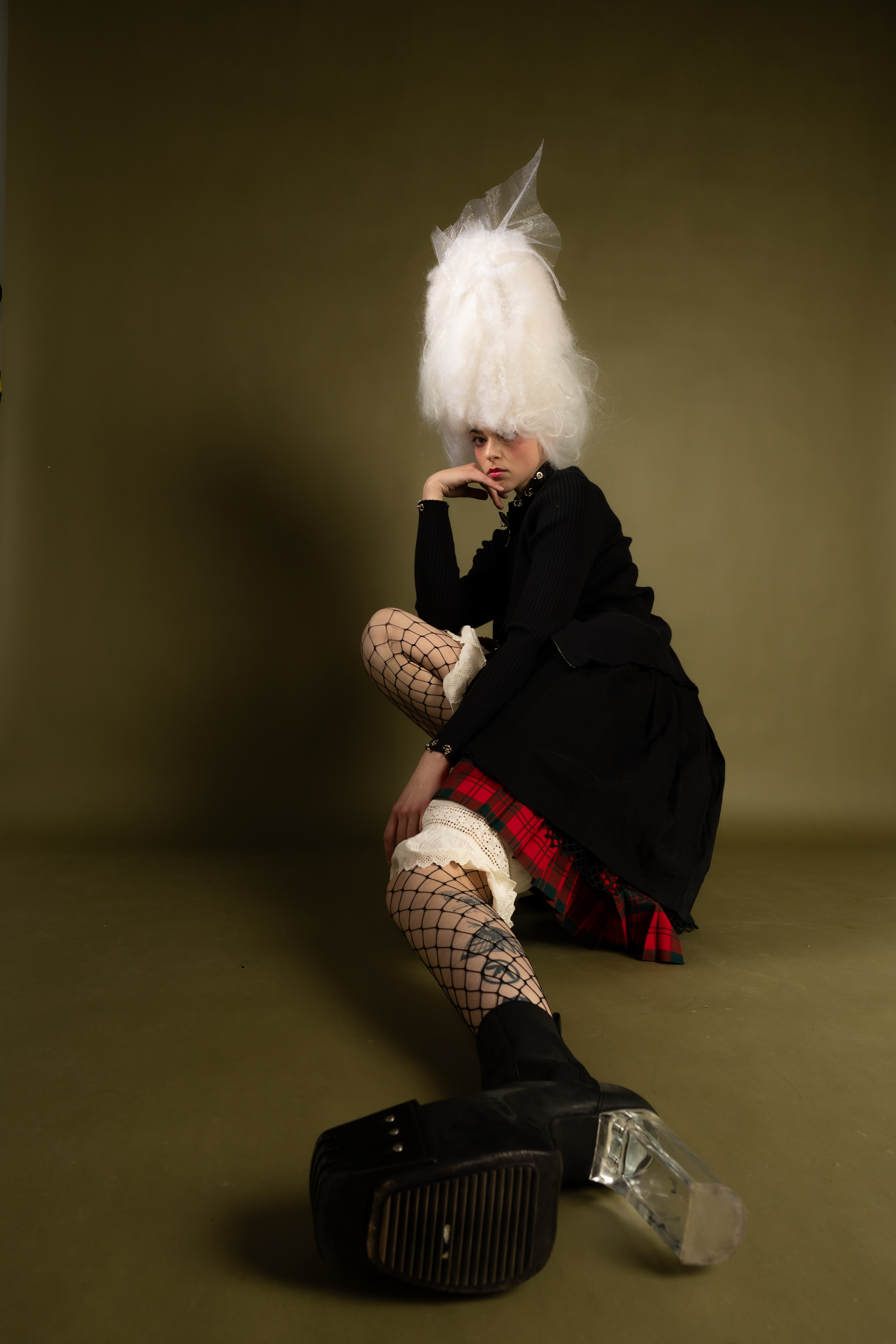 Model in voluminous white headpiece and black dress wearing fishnet tights and platform shoes against a dark olive backdrop.