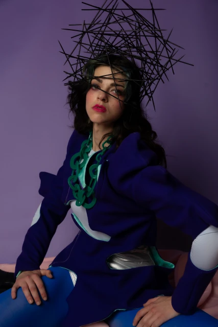 A woman in a blue futuristic costume with a black headpiece sits against a purple background, creating a fashion-forward, artistic image.