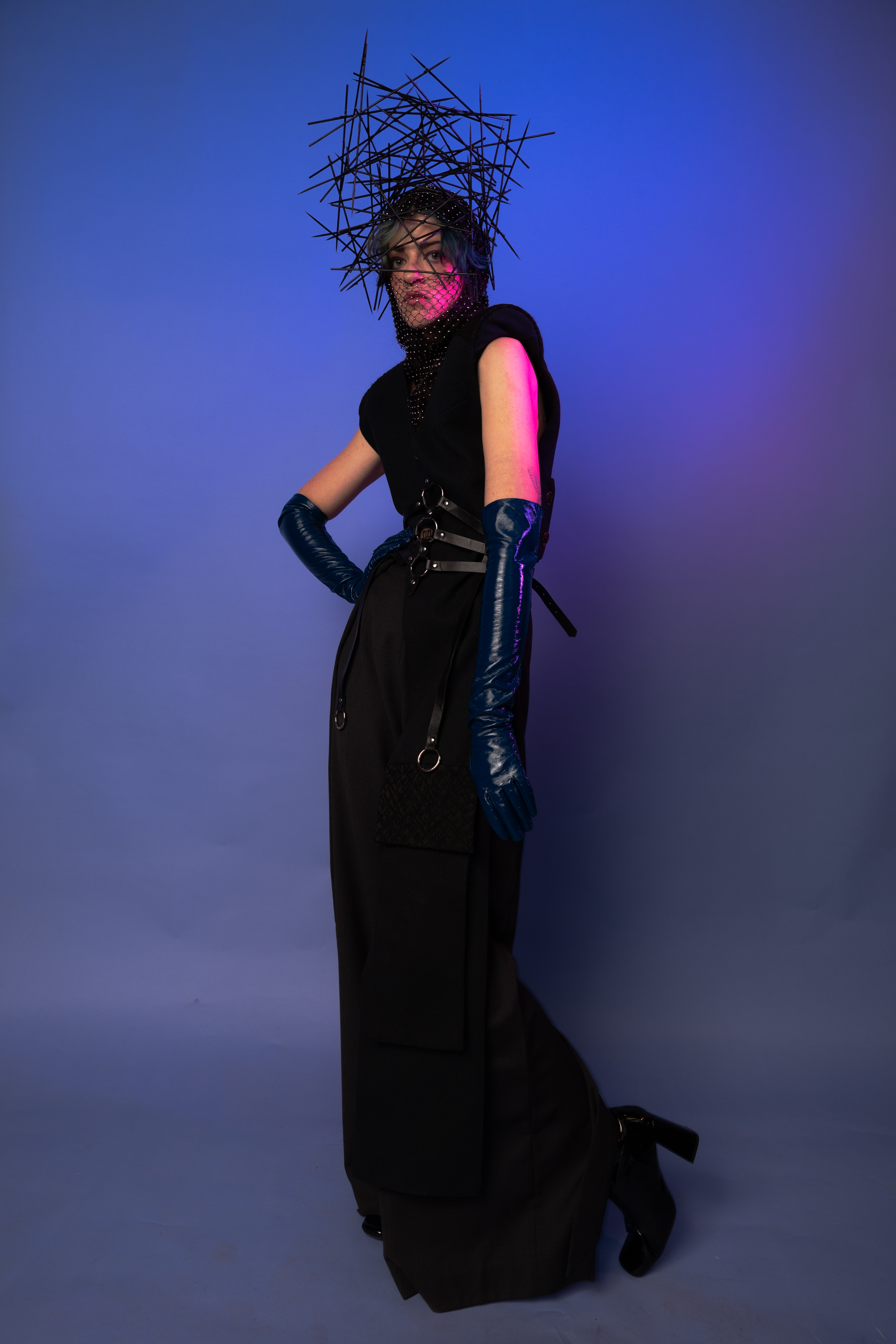 Full-length shot of a person in a dark outfit and blue gloves, wearing a dark headdress, posing against a blue and purple backdrop.