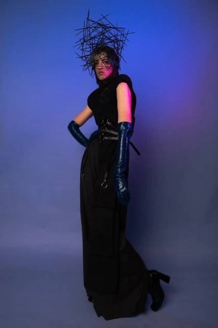 Full-length shot of a person in a dark outfit and blue gloves, wearing a dark headdress, posing against a blue and purple backdrop.