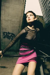 Model with short dark hair wearing a pink miniskirt and mesh top posing outdoors against an urban concrete wall.