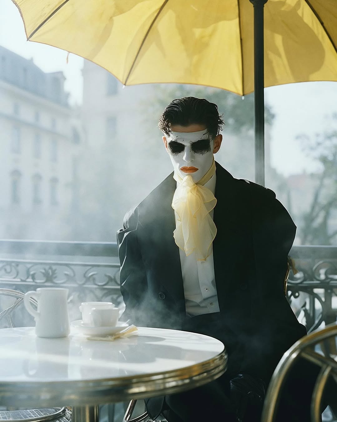Person with white face makeup in black suit sitting at outdoor cafe table beneath yellow umbrella in urban environment with white building.