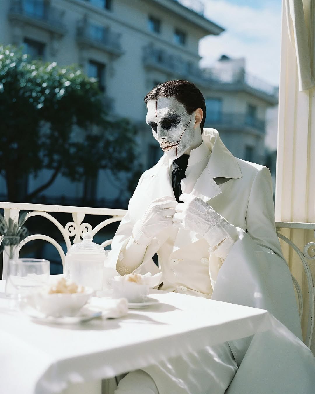 Person with theatrical makeup and a white suit sitting at a table with teacups outside a building and trees on a sunny day.