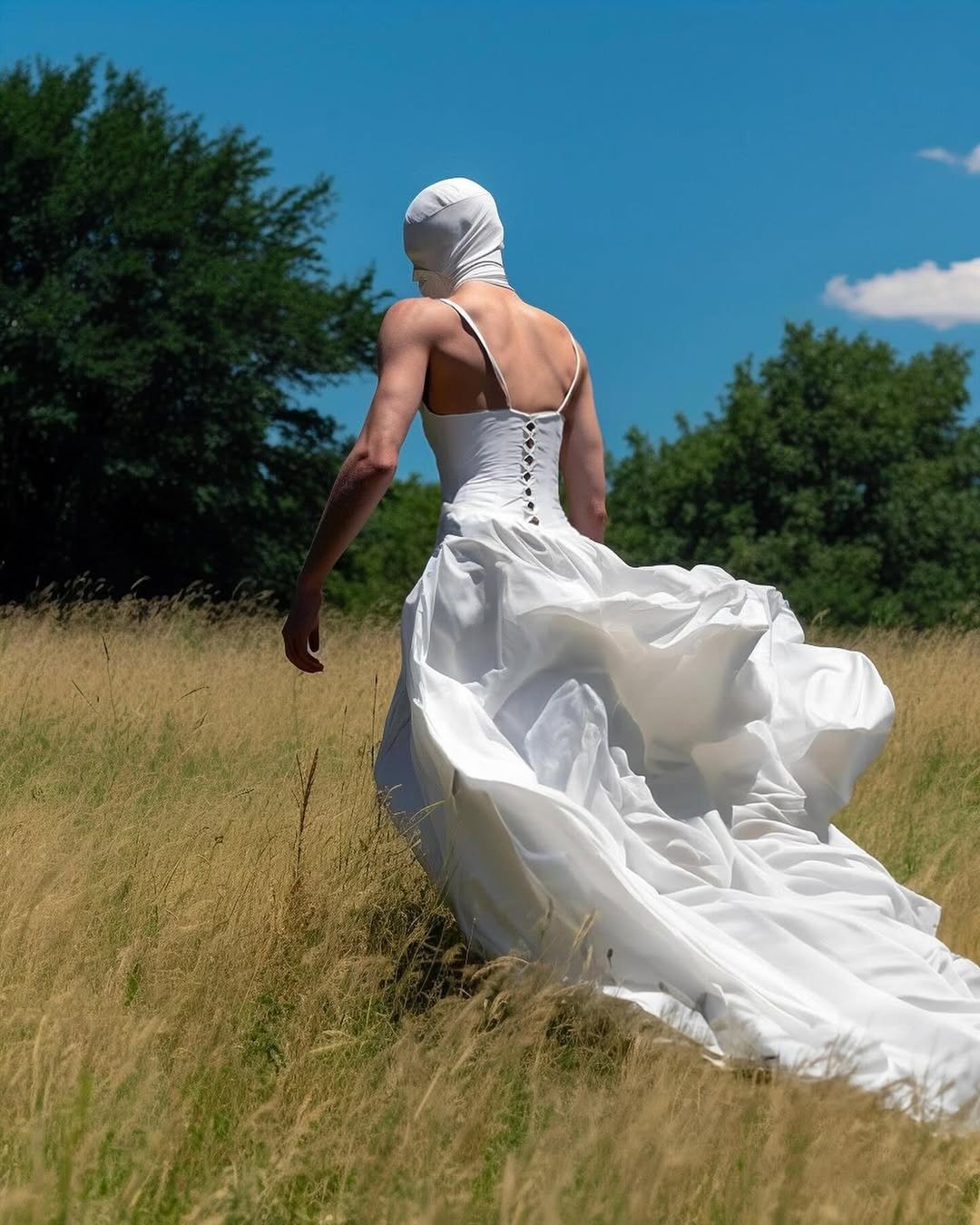 Person in a white dress and head covering walking through a grassy field on a sunny day with a blue sky and trees in the background.