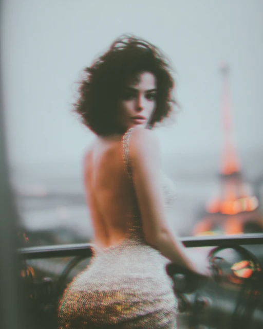 A woman in a glittering dress poses on a balcony overlooking the Eiffel Tower in Paris. The image uses a shallow depth of field.