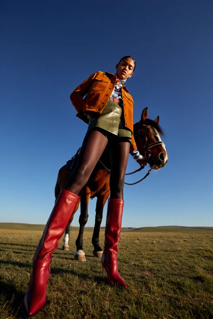 A woman in riding boots poses on horseback in a grassy field under a clear blue sky, showcasing high fashion in a rural setting.