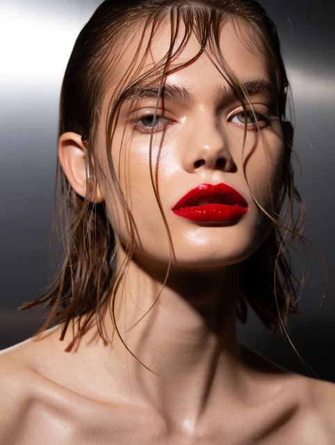 Close-up portrait of a young woman with wet hair and bright red lipstick, against a dark gray background. Beauty shot with vibrant makeup.