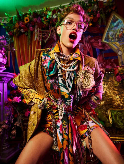 Eccentric portrait: Person with brown hair, glasses, gold coat, floral top, and chain belt poses in front of flower backdrop.